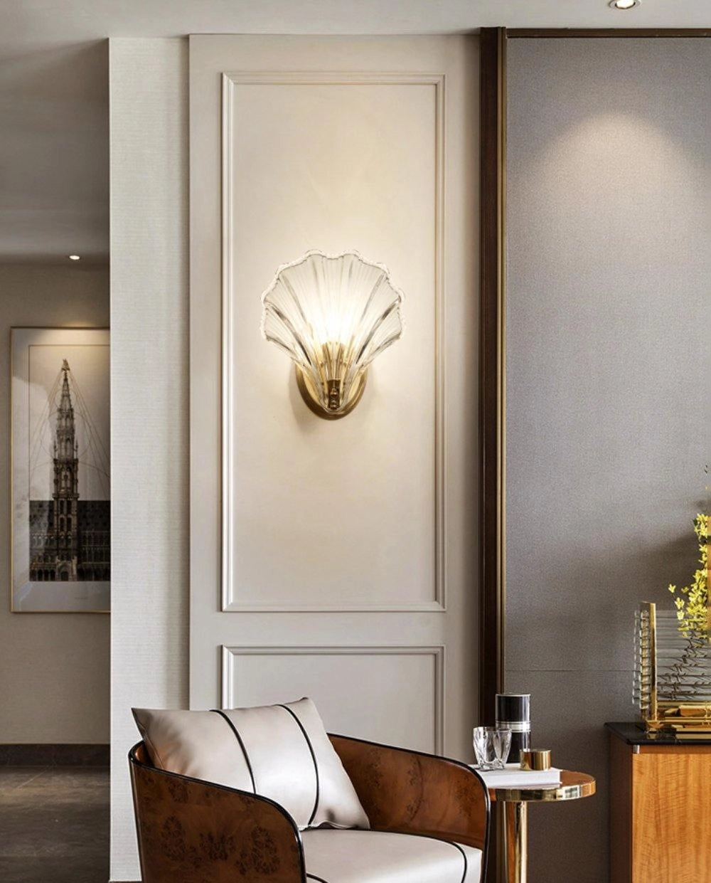 Gold nautical coastal seashell wall sconce light fixture mounted on a white wall in a living room with wood flooring and picture frames.