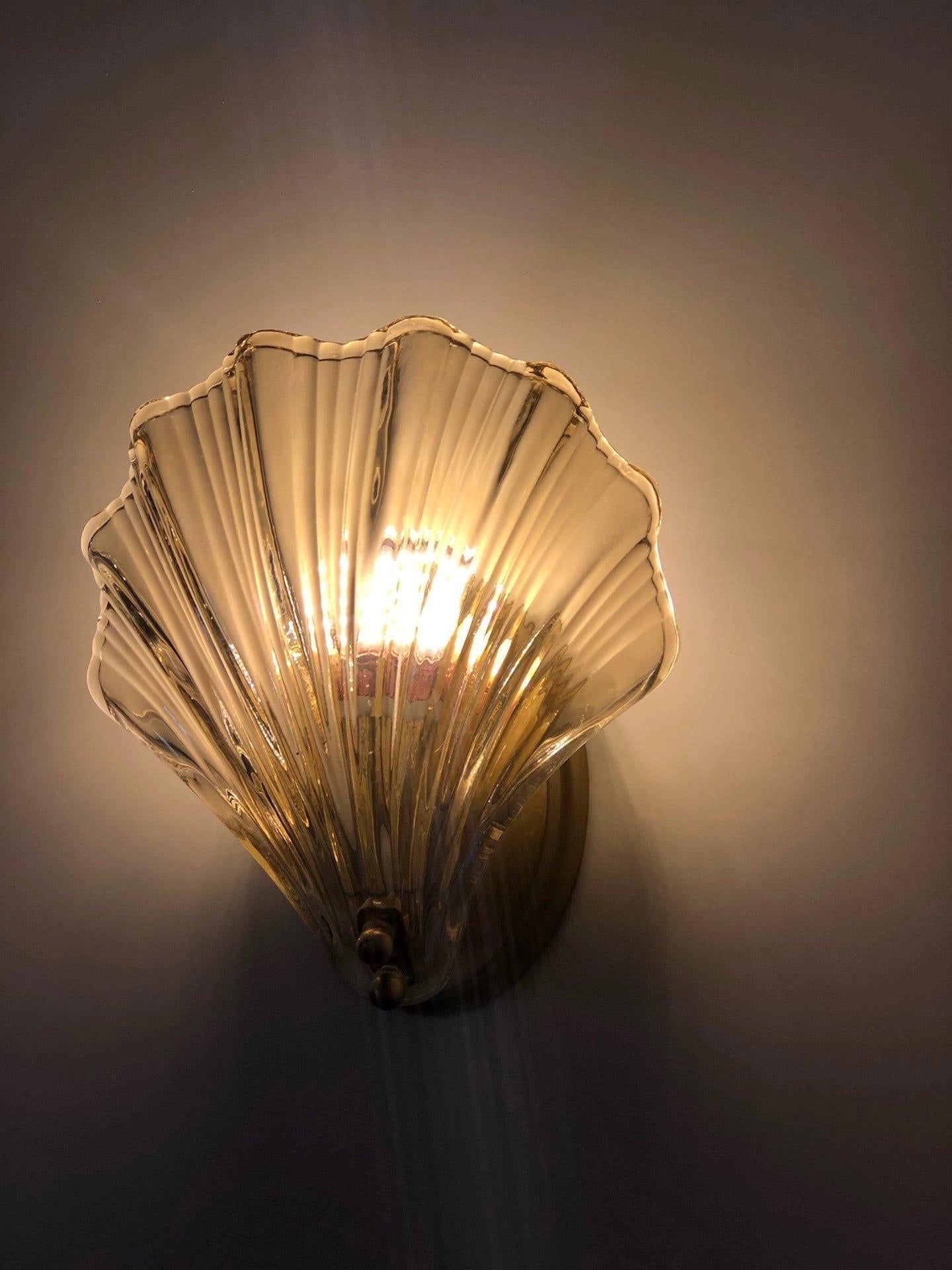 Gold nautical coastal seashell wall sconce light fixture mounted on a wooden surface, illuminating the area, with a sky and tree in the background.