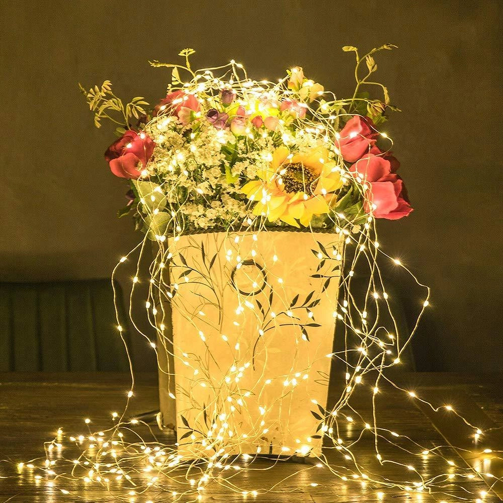 Solar powered outdoor LED fairy lights wrapped around a plant with decorative flower arrangements in mason jars, creating a warm amber glow suitable for garden or patio decoration.