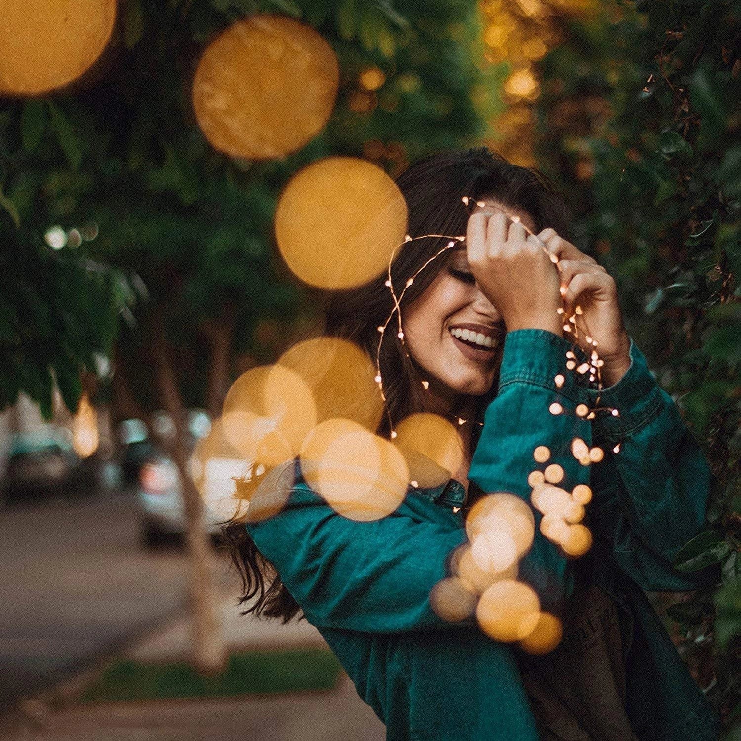 Outdoor setting with solar powered LED fairy lights illuminating a smiling person with black hair during summer, surrounded by trees.