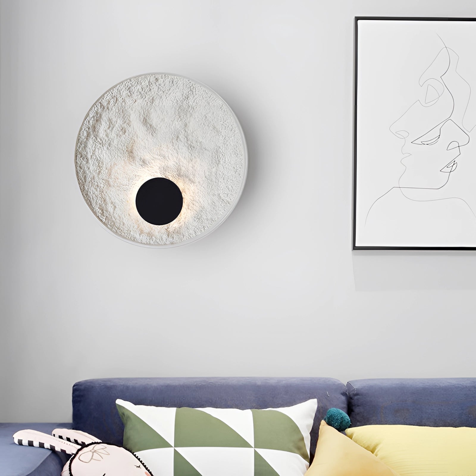 Minimalist contemporary circular wall sconce with textured design mounted on a grey wall, complementing a modern interior with white and black accents, including a grey couch.