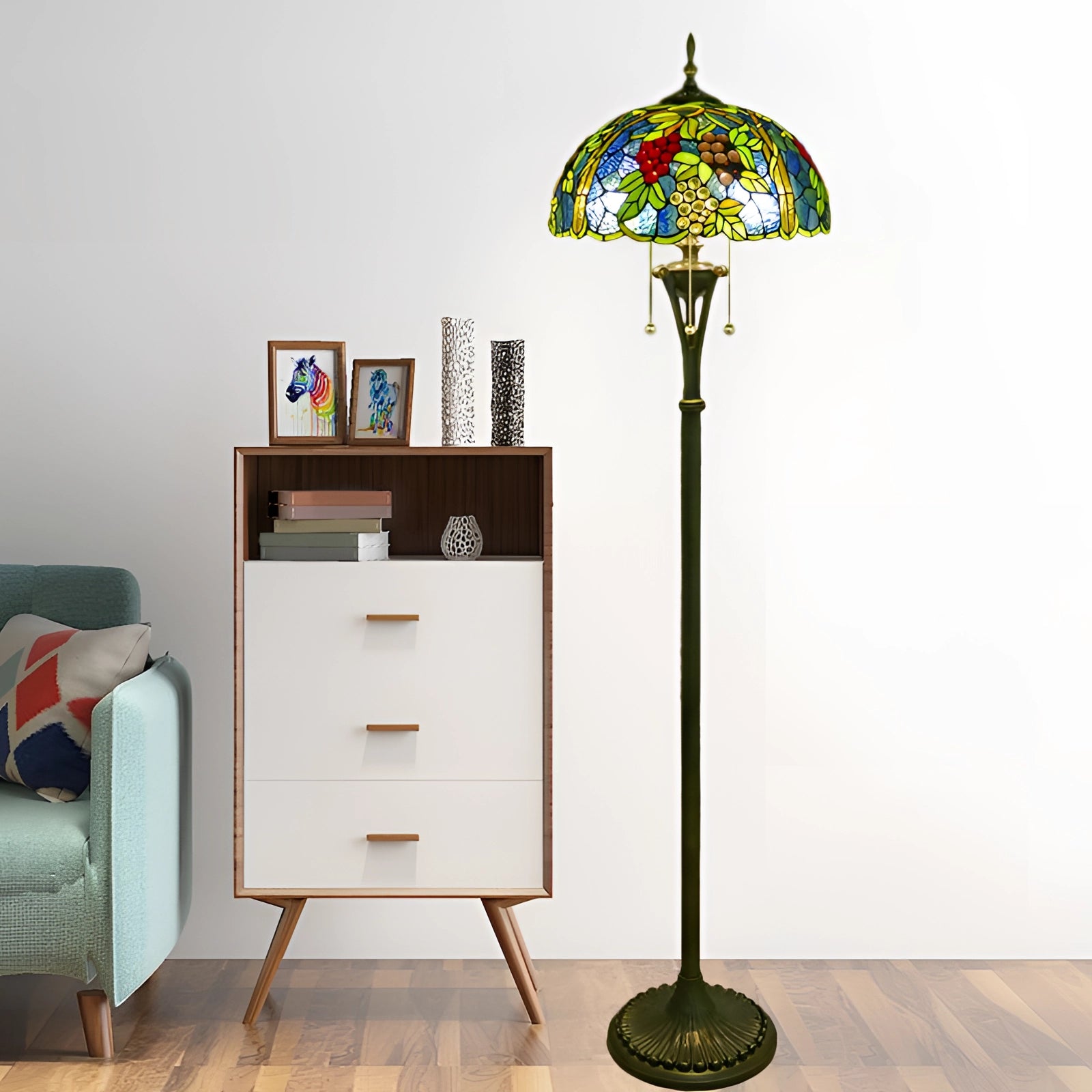 Tiffany-style floor lamp with a stained glass peacock feather design, featuring a wooden base and standing beside a wooden cabinet with drawers and shelving, amidst a room setting with a plant and wooden flooring.