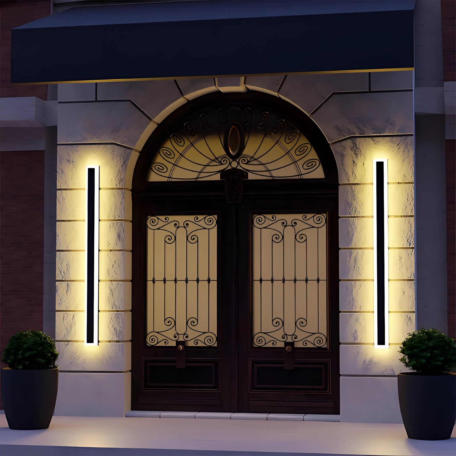 Vertical LED wall light illuminating an outdoor setting with a minimalist design, mounted near a door and window on a wooden exterior wall, surrounded by potted plants and greenery.