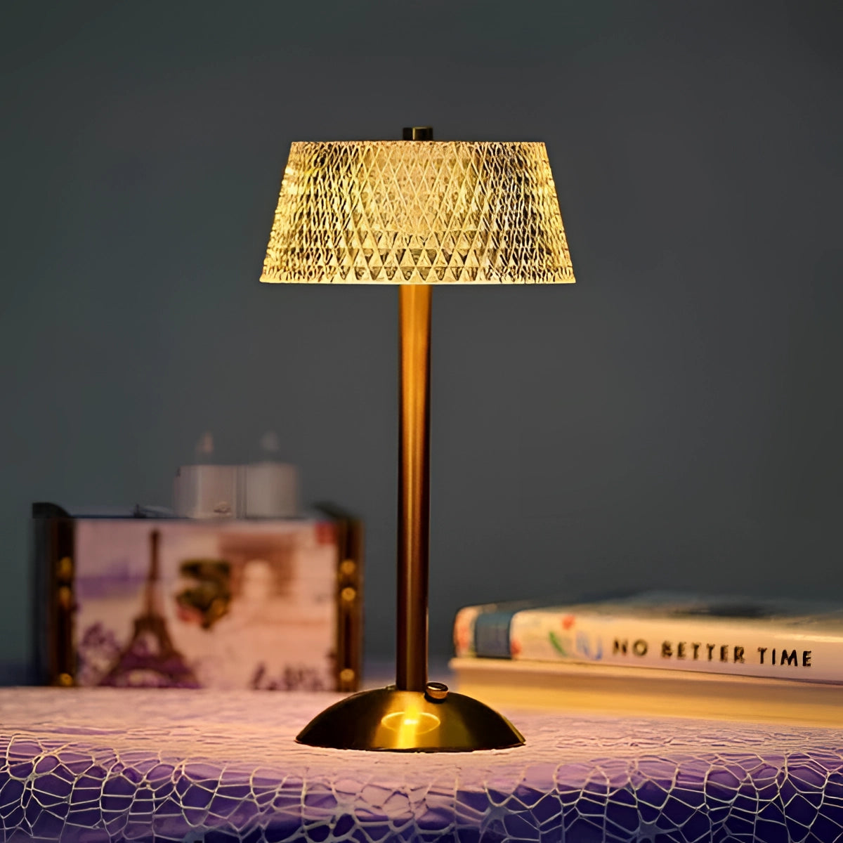 Vintage brass table lamp with pleated shade placed on a wooden surface in a dimly lit room, accompanied by books and artistic decor.