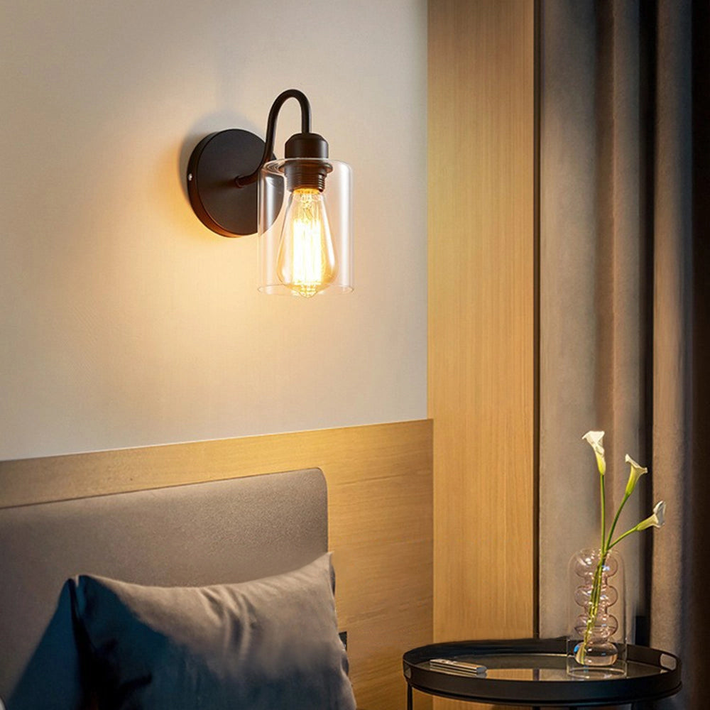 Vintage Edison bulb wall sconce with black metal frame and clear glass shade, mounted on a wooden wall in a cozy interior setting, illuminating the space with warm yellow light.