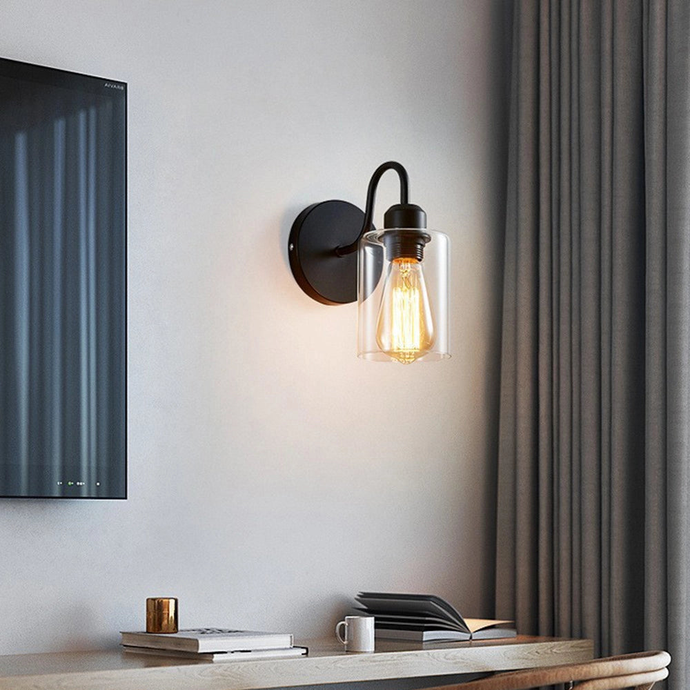 Vintage Edison bulb wall sconce featuring a black metal frame and clear glass shade, mounted on a wall in an interior space with wooden accents and a window with curtains.