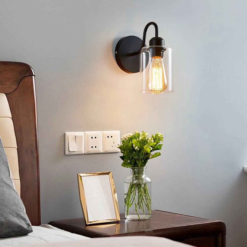 Vintage Edison bulb wall sconce with a black metal frame and clear glass, mounted on a wall in an interior setting. The room features wooden elements and a cozy ambiance with a table and floor visible.