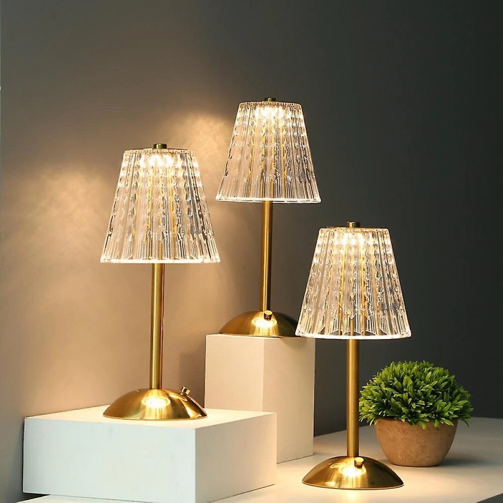 Vintage gold base pleated table lamp with soft lighting, placed on a wooden surface beside a potted plant, set against a warmly colored interior design scene.