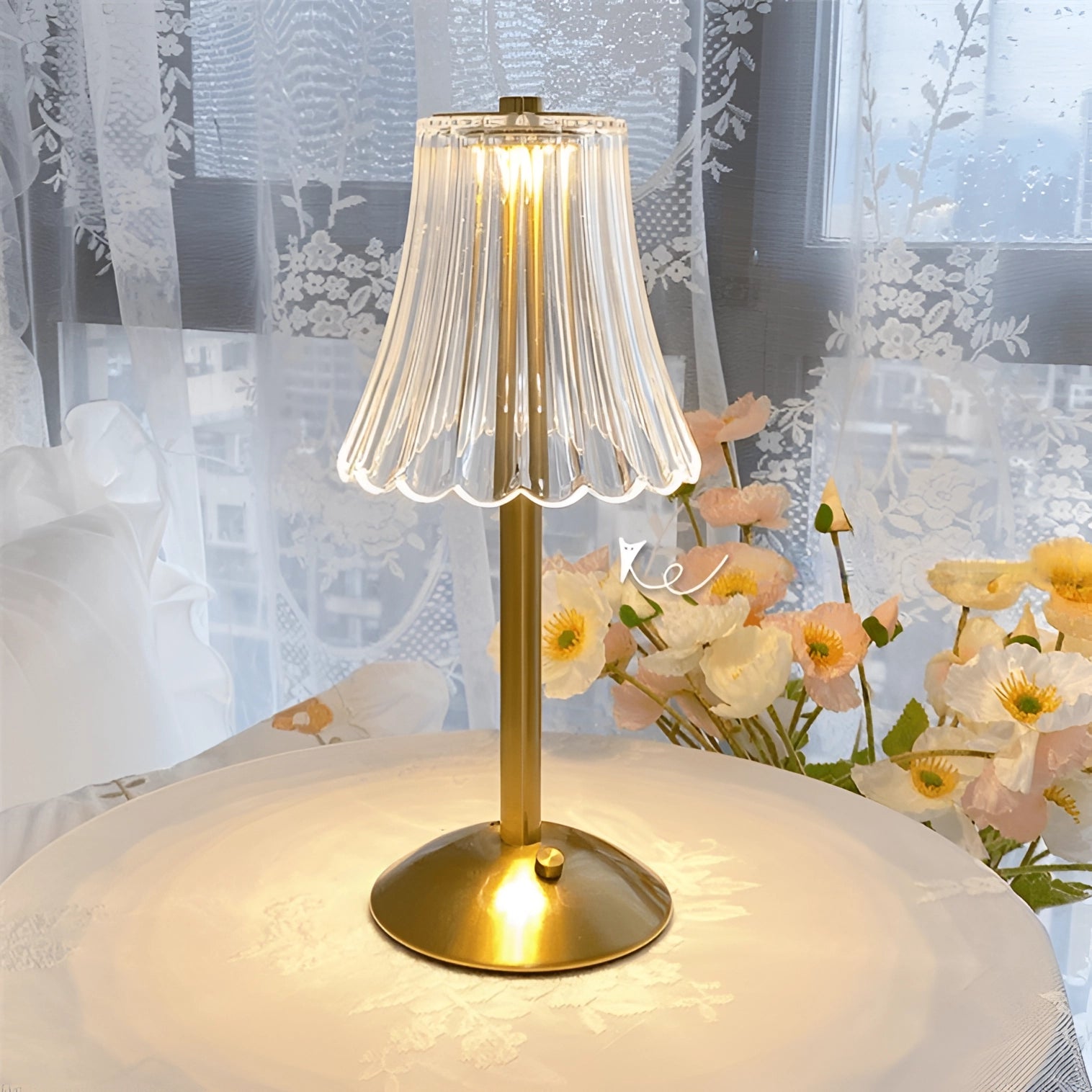 Vintage gold base pleated table lamp with soft lighting, placed on a wooden table, accompanied by a small green plant and decorative art pieces in a stylishly designed interior space.