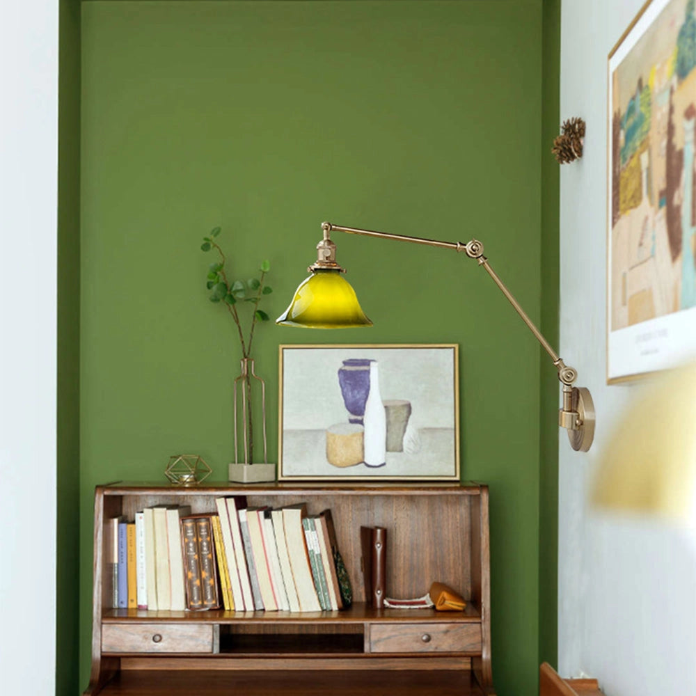 Vintage green enamel wall lamp with a brass arm mounted on the wall above a wooden shelf, next to a picture frame, houseplant, and bookcase in a stylish interior design setting.