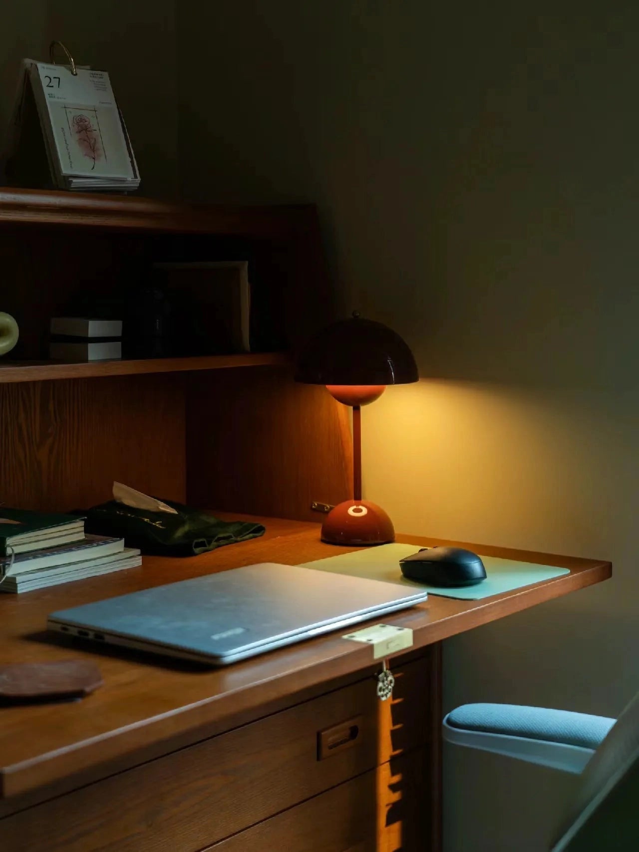 Vintage red desk lamp with a retro design, featuring a wooden base and arm, placed on a wooden shelf in a home office setting. The lamp is turned on, casting a warm light in a room with wooden cabinetry and modern interior design elements.