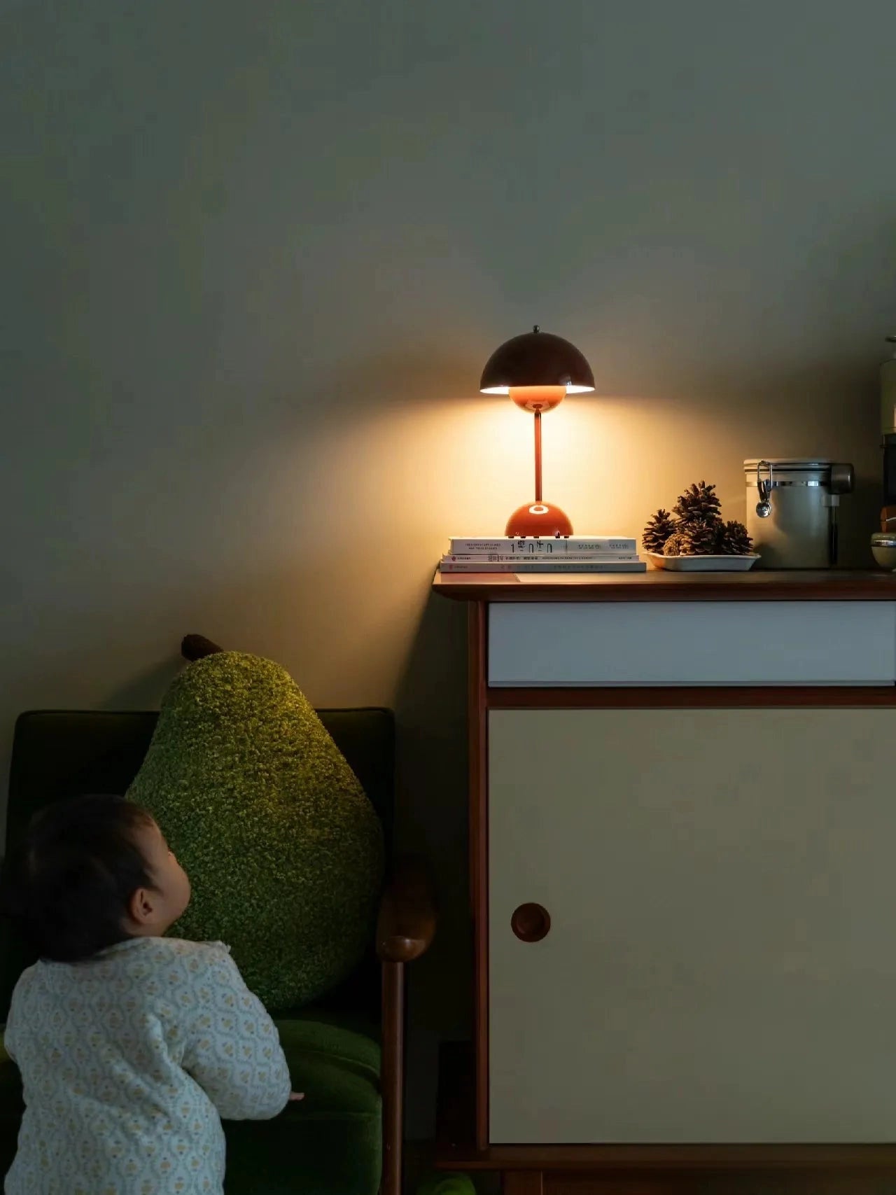 Vintage red desk lamp with a wooden base, emitting a warm yellow light, placed on a table against a wall, in a cozy room setting with a wooden floor and furniture featuring drawers.