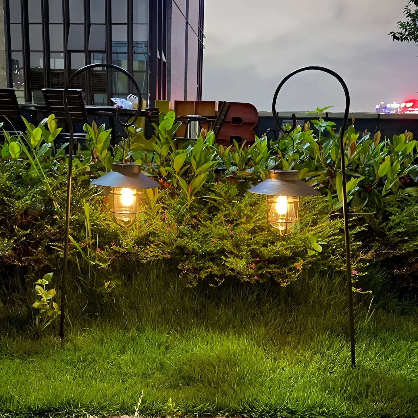 Vintage Solar LED Garden Lantern illuminating a garden area with surrounding plants and vegetation, set against a backdrop of a clear sky and nearby buildings.