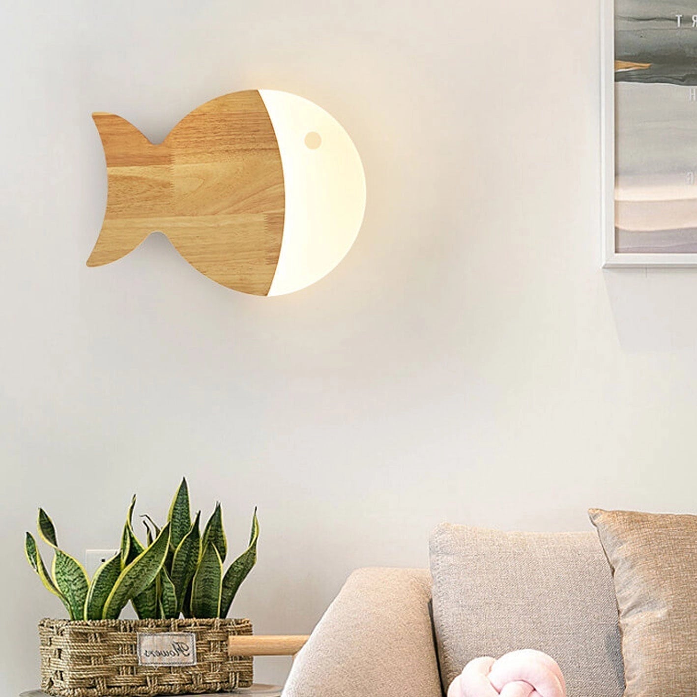 Wooden Fish LED Wall Light mounted on a wall, featuring a nautical and Scandinavian design, with a wooden finish. The setting includes cozy interior elements like a couch and a terrestrial plant, creating a comfortable and stylish atmosphere.