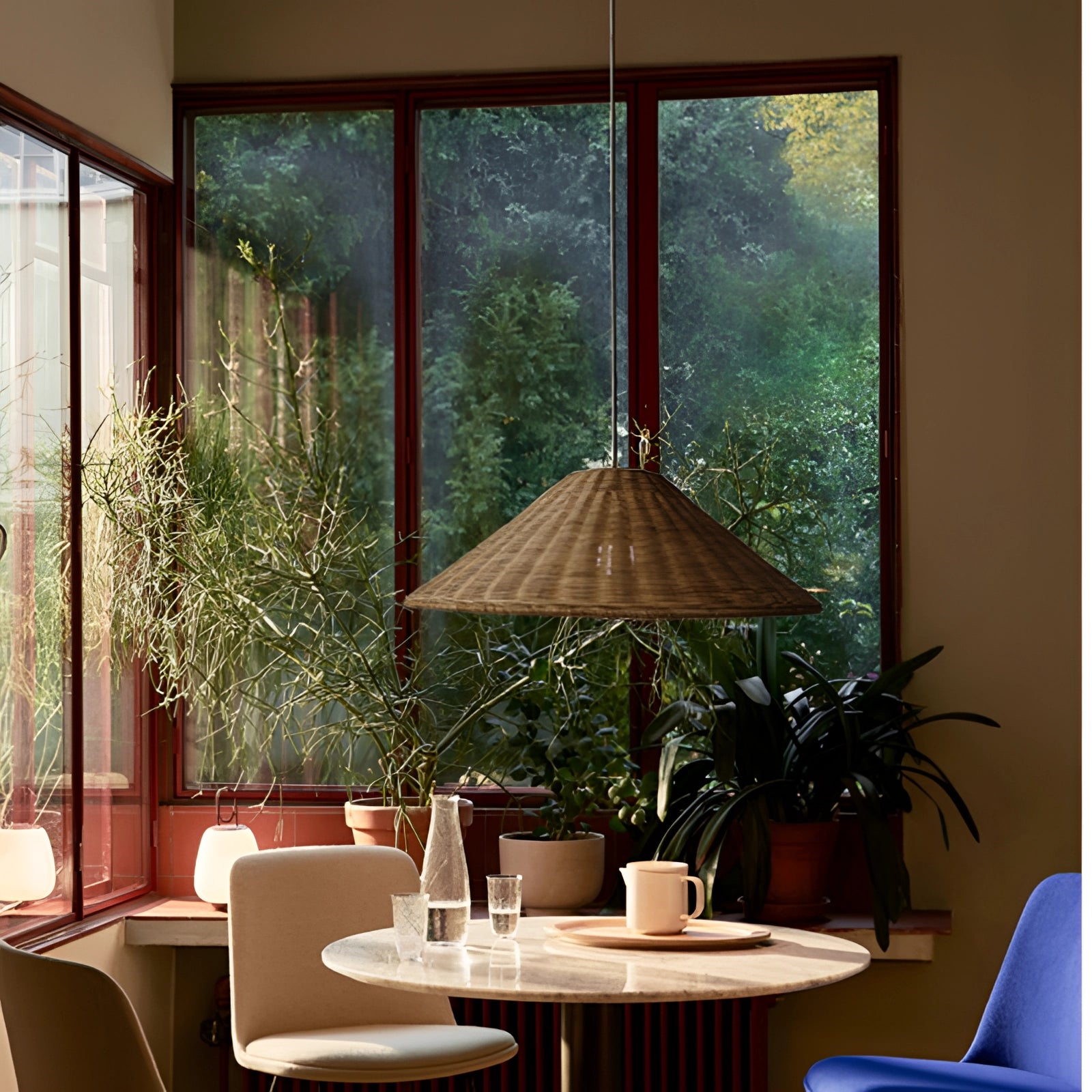 Eco-friendly woven rattan pendant light with a conical shape, hanging in a Scandinavian-style interior featuring a wooden table, potted plant, large window, and minimalist furniture.